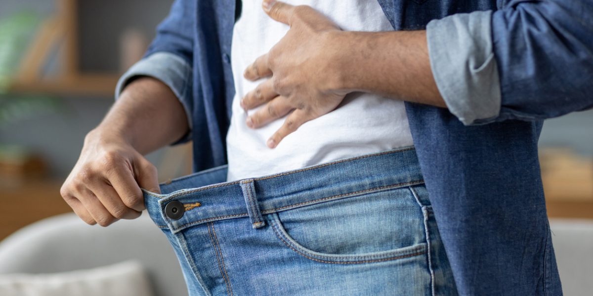 A man shows how much weight he has lost by holding out the waist of his jeans, symbolizing his successful diet.