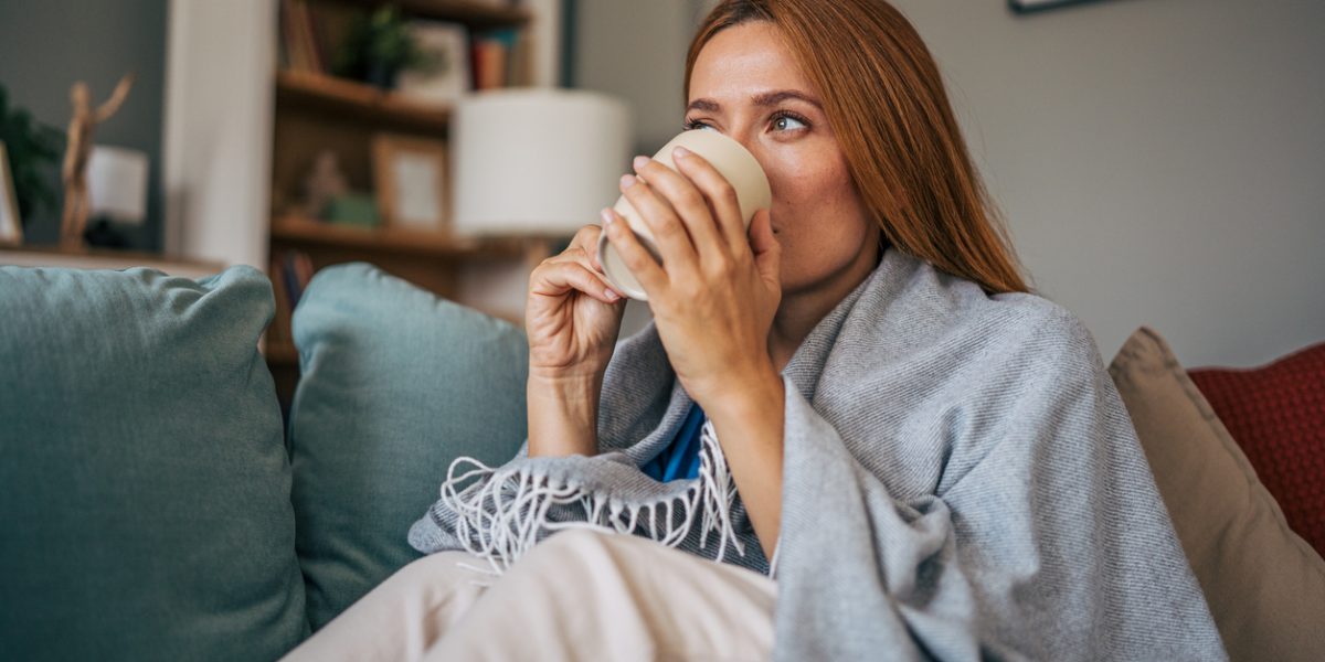 Cozy portrait of a sick mid adult woman wrapped in a blanket, enjoying a warm drink on the sofa in her living room, finding comfort and relief