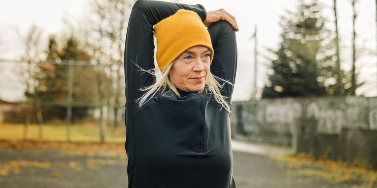 Mature woman stretching arms in the city park. Female jogger wearing knitted hat doing warm up workout outdoor on a winter morning.